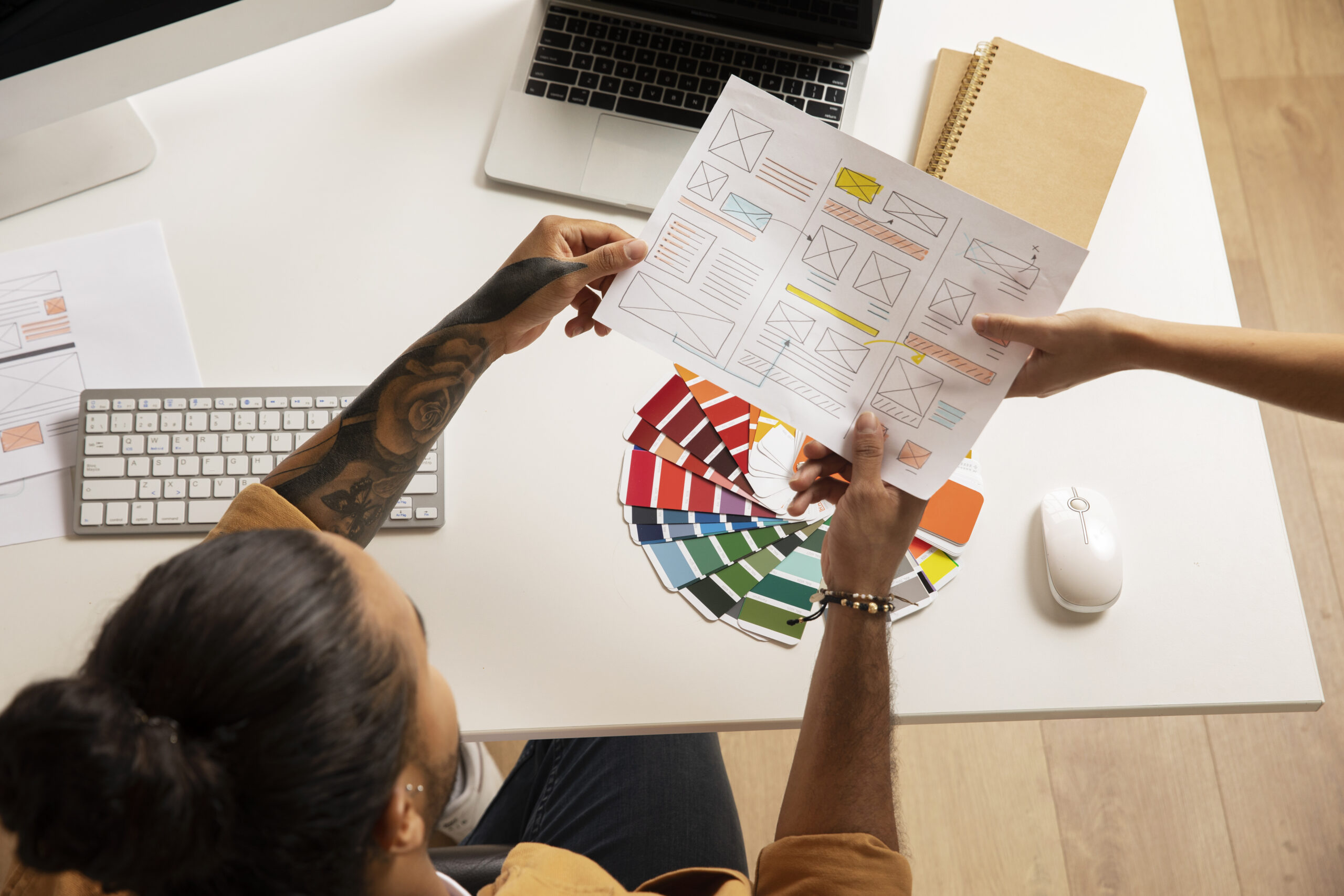 Designer segurando protótipos de layout e paleta de cores em frente ao computador, em ambiente de trabalho criativo.
