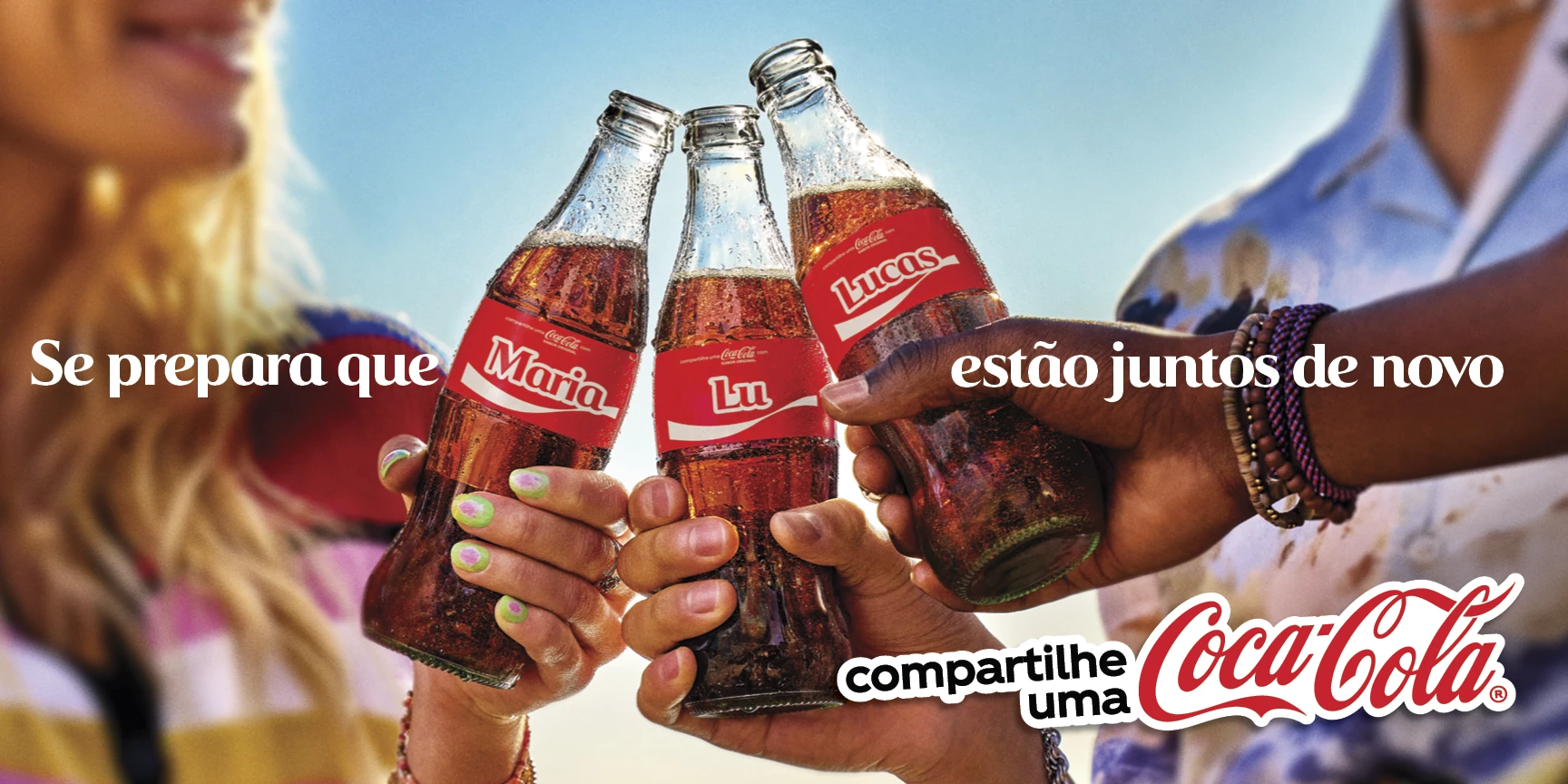 Três pessoas brindando com garrafas de Coca-Cola personalizadas com os nomes Maria, Lu e Lucas, celebrando o retorno da campanha ‘Compartilhe uma Coca-Cola